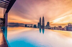 Une vue urbaine offrant une perspective dégagée sur les tours jumelles Petronas, qui se reflètent dans une étendue d’eau calme, probablement une piscine, avec un bâtiment moderne sur la gauche.
