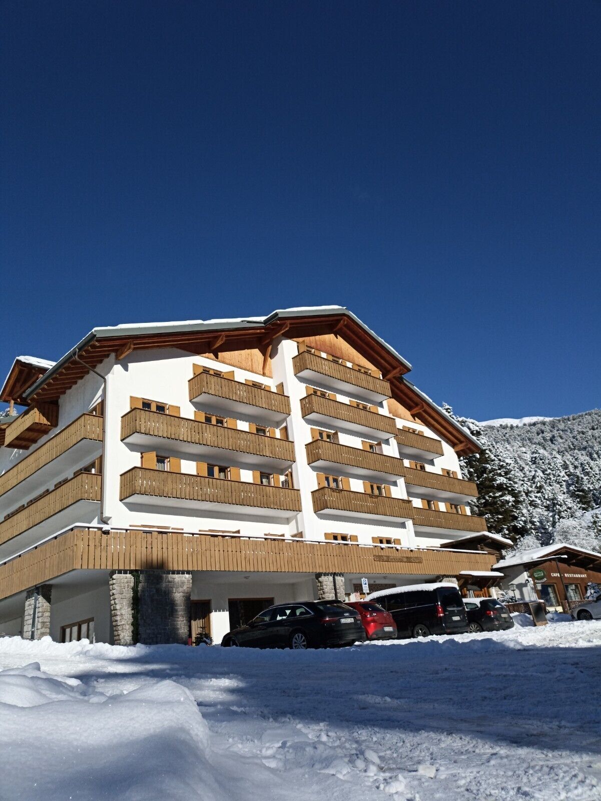 A multi-story hotel with wooden balconies, a ski slope in the background, and a clear blue sky.