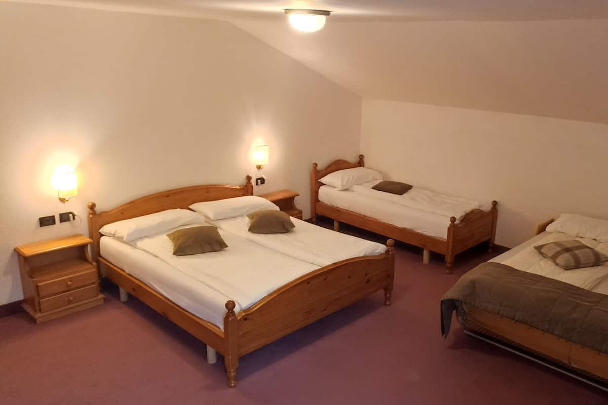 Habitación de hotel con tres camas de madera, cada una con ropa blanca y almohadas marrones. Hay mesitas de noche con lámparas y un cajón pequeño. El techo es inclinado y tiene un accesorio de iluminación.