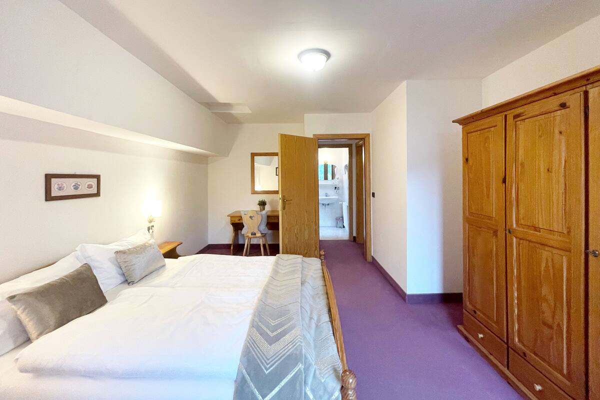 Un dormitorio con una cama grande, un armario de madera, un escritorio con una silla, y un baño visible a través de una puerta abierta.
