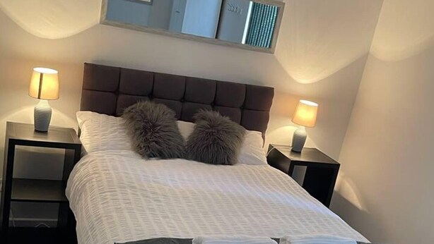 A neatly made bed with a headboard, two bedside lamps, and a mirror above the bed. - Modern and Cosy Apartment in Leeds, England (Leeds)