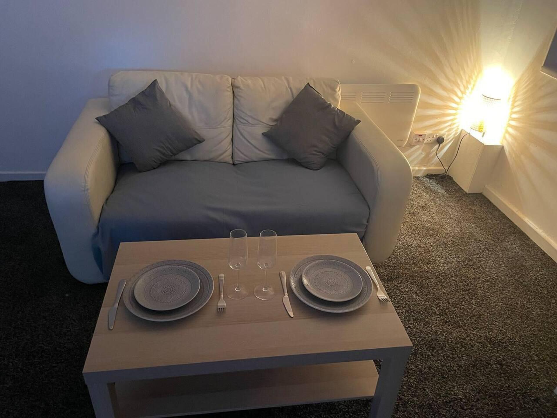 A dining setup with a table, plates, cutlery, and glasses, positioned in front of a sofa with cushions.