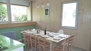 A dining area with a wooden table set for four, surrounded by chairs. There are windows providing natural light, and a small picture frame on the wall.