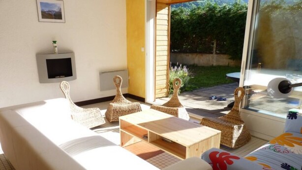 A sunlit room with a sofa, two woven poufs, a wooden coffee table, and a window with a view of a garden.