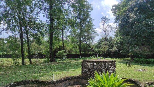 A park with a stone wall, a bench, and a circular concrete planter with plants. - Homecation Manas View Jungle Resort (Barnagar)
