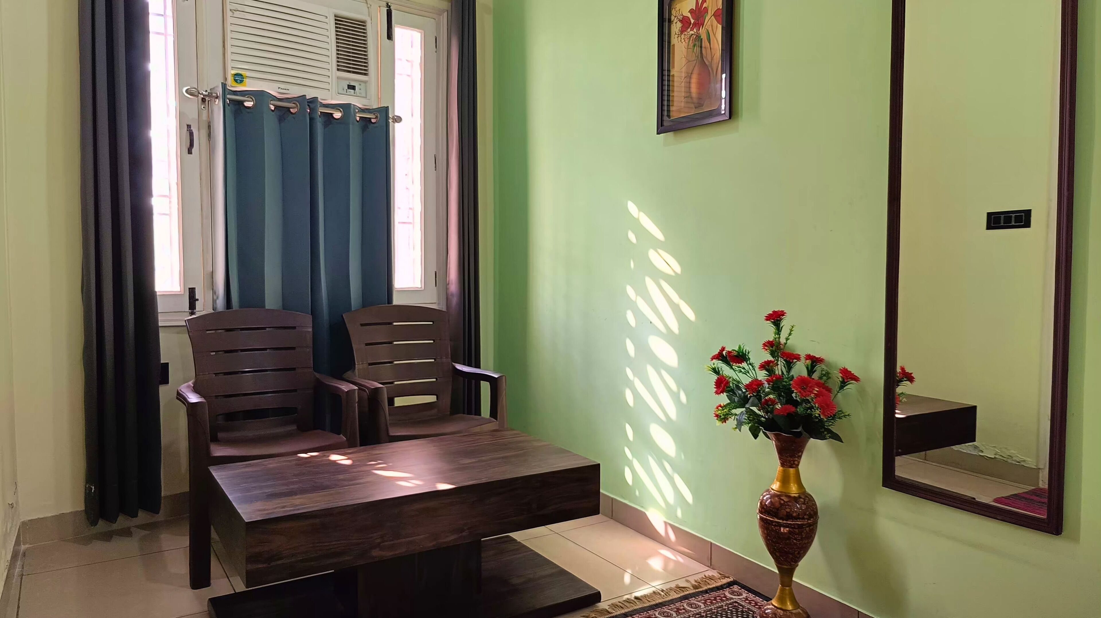A room with two wooden chairs, a dark wooden table, a vase with red flowers, a mirror, and a window with blue curtains.