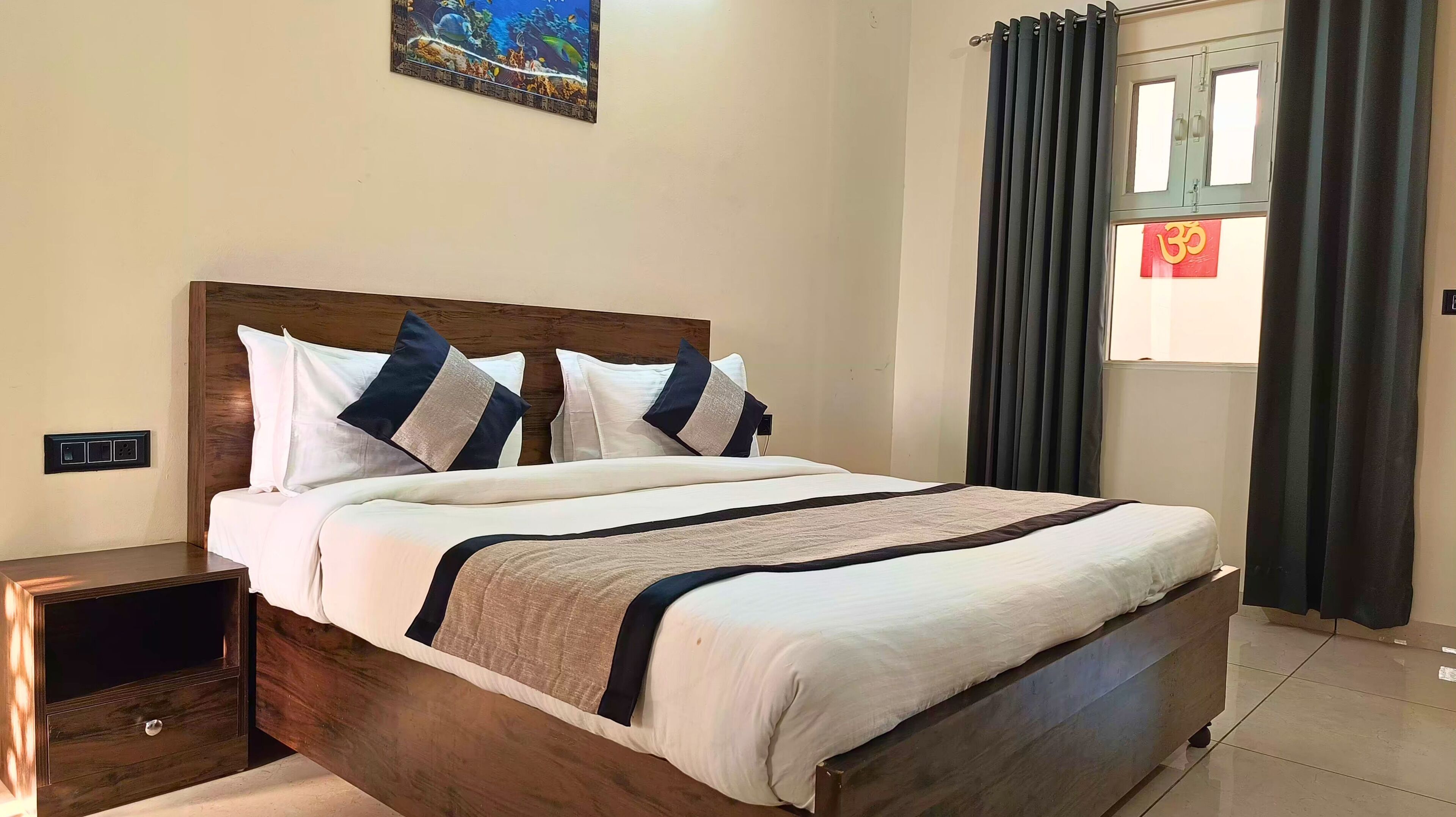 A neatly made bed with a wooden headboard, white and dark blue pillows, and a beige and dark blue bedspread. There is a nightstand with a drawer, a wall-mounted power outlet, and a window with a curtain.