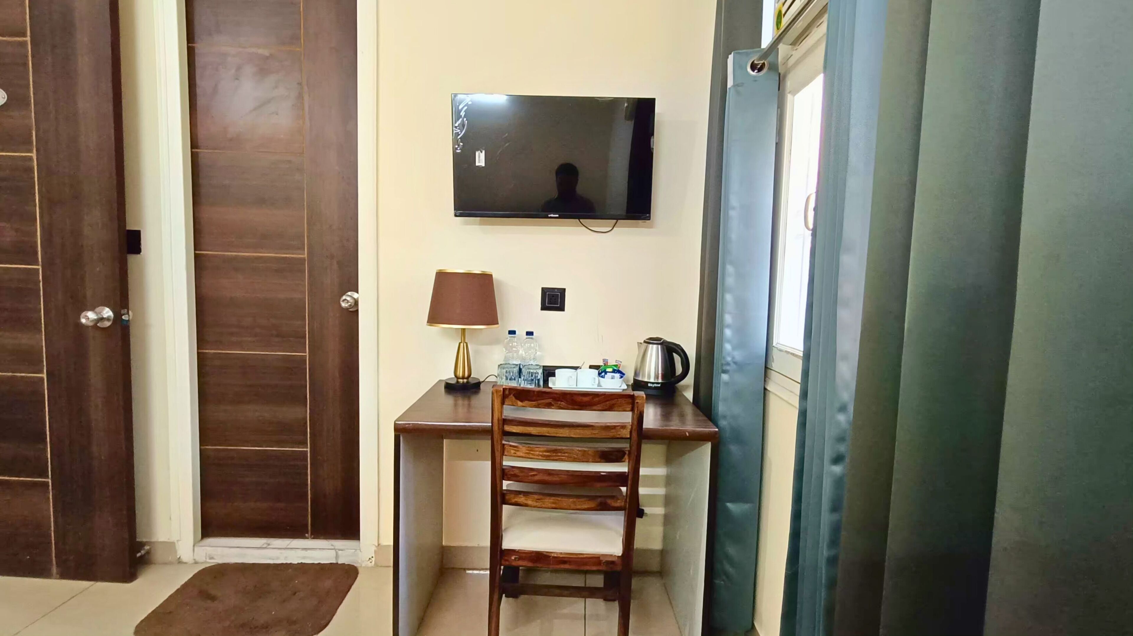 A small room with a wooden desk, a chair, a television mounted on the wall, a lamp, and a window with curtains.