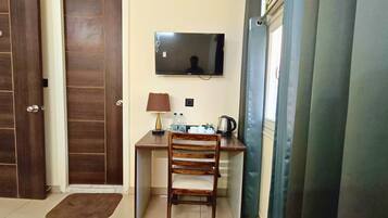 Una habitación pequeña con un escritorio de madera, una silla, un televisor montado en la pared, una lámpara y un ventanal con cortinas.