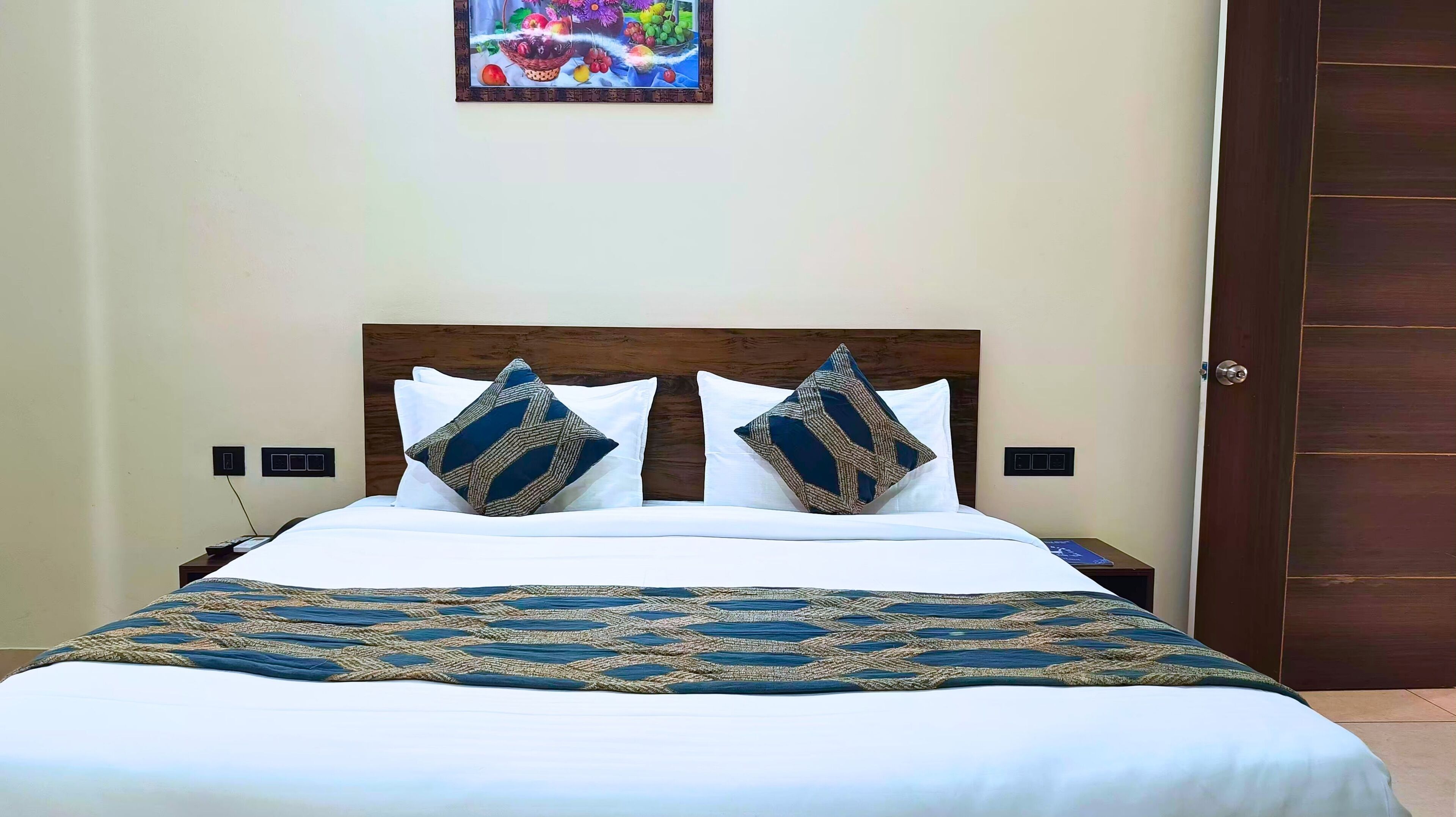 Een net opgemaakt bed met een bedsprei met patroon en twee decoratieve kussens. Daarboven hangt een klein schilderij aan de muur.