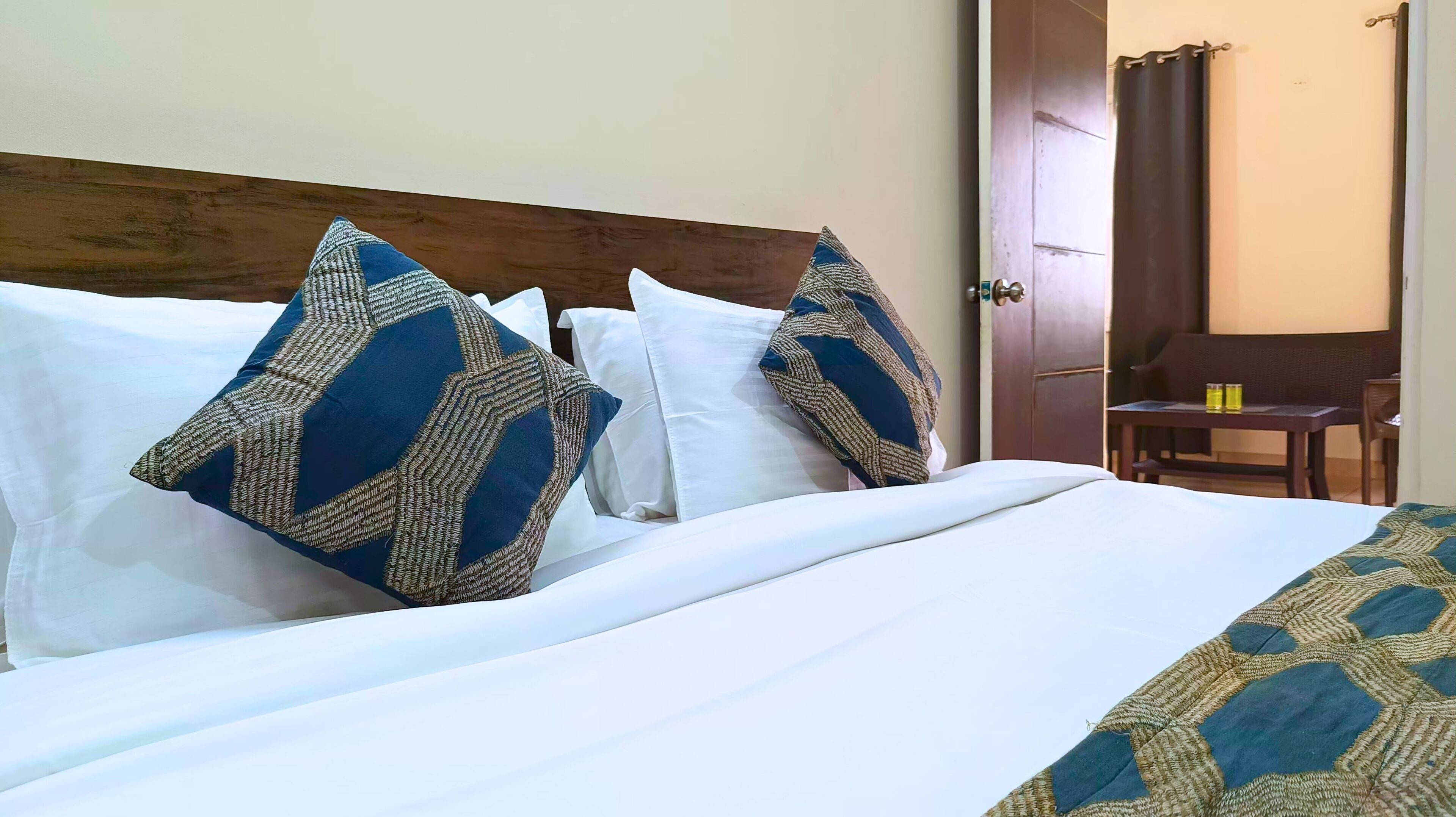 A neatly made bed with white linens and patterned pillows, a wooden headboard, and a small table with two yellow bottles in the background.
