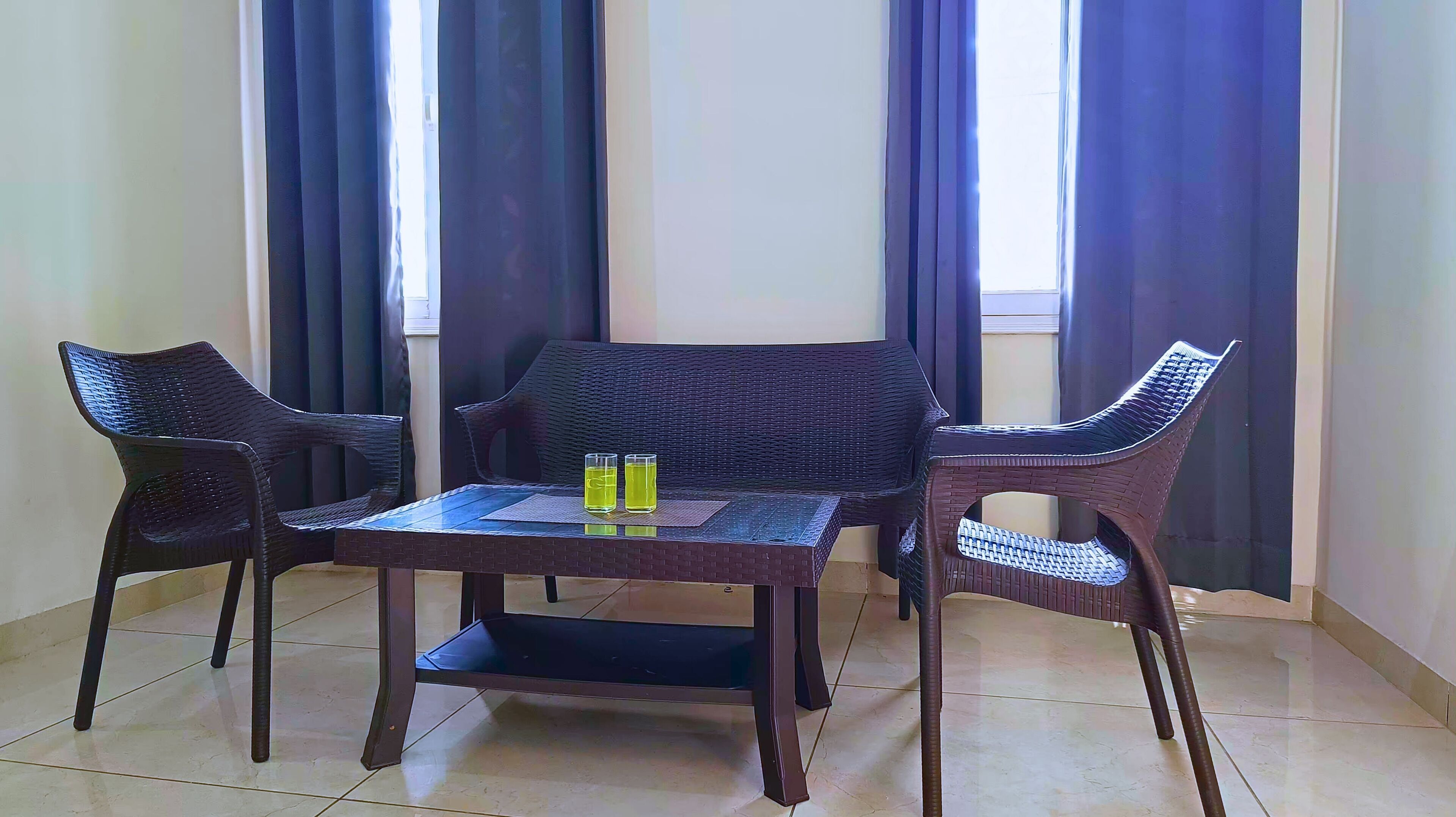 Una habitación con una mesa y sillas de mimbre oscuro, dos vasos de líquido amarillo sobre la mesa, y una vista a través del ventanal.