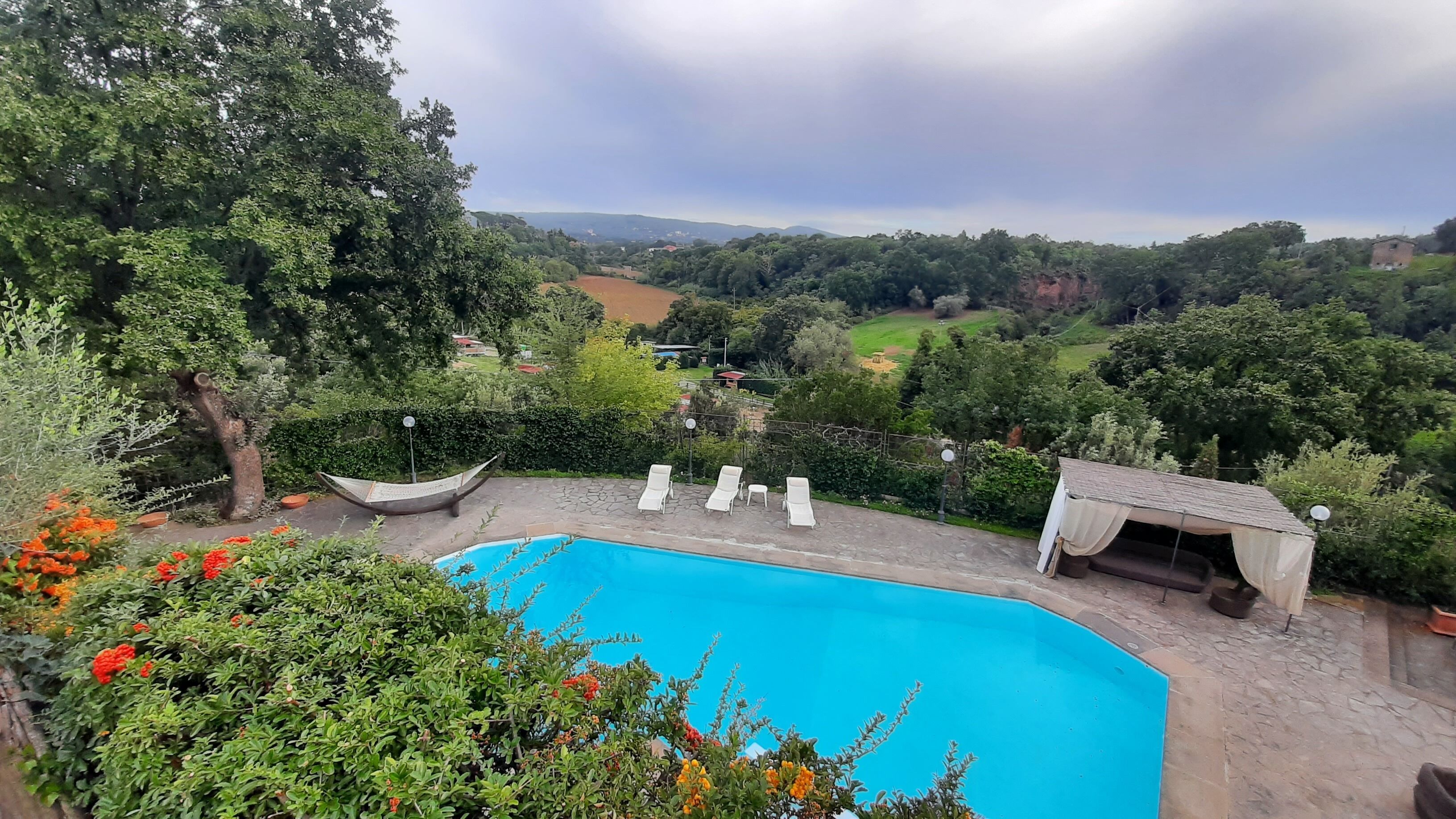 Uma piscina com espreguiçadeiras, rodeada por vegetação exuberante e vista para colinas ao longe.