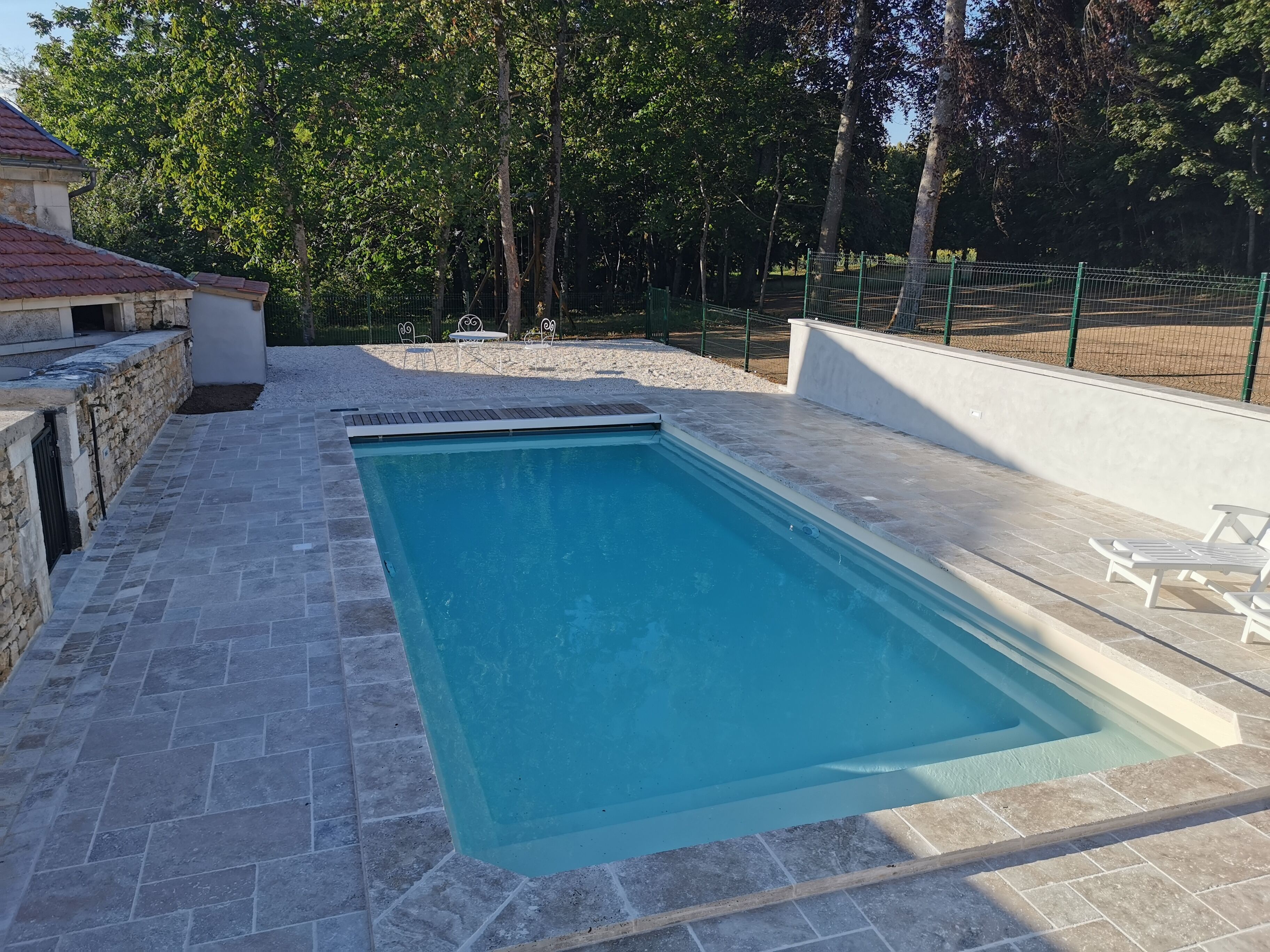 A rectangular swimming pool with a stone border, surrounded by a white wall and a green fence.