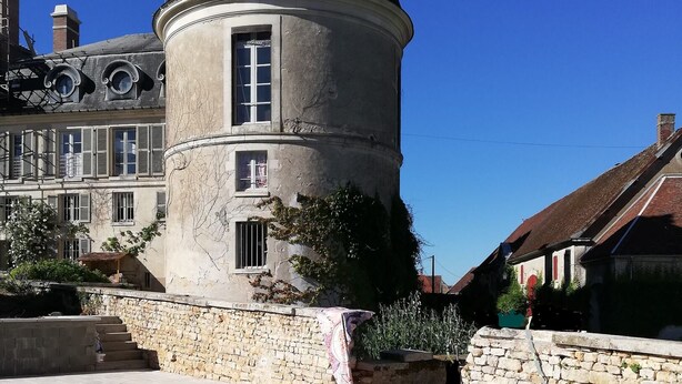 Une piscine bordée d’un mur en pierre et dotée d’une terrasse en bois, entourée d’un bâtiment historique avec une tour ronde et de nombreuses fenêtres.