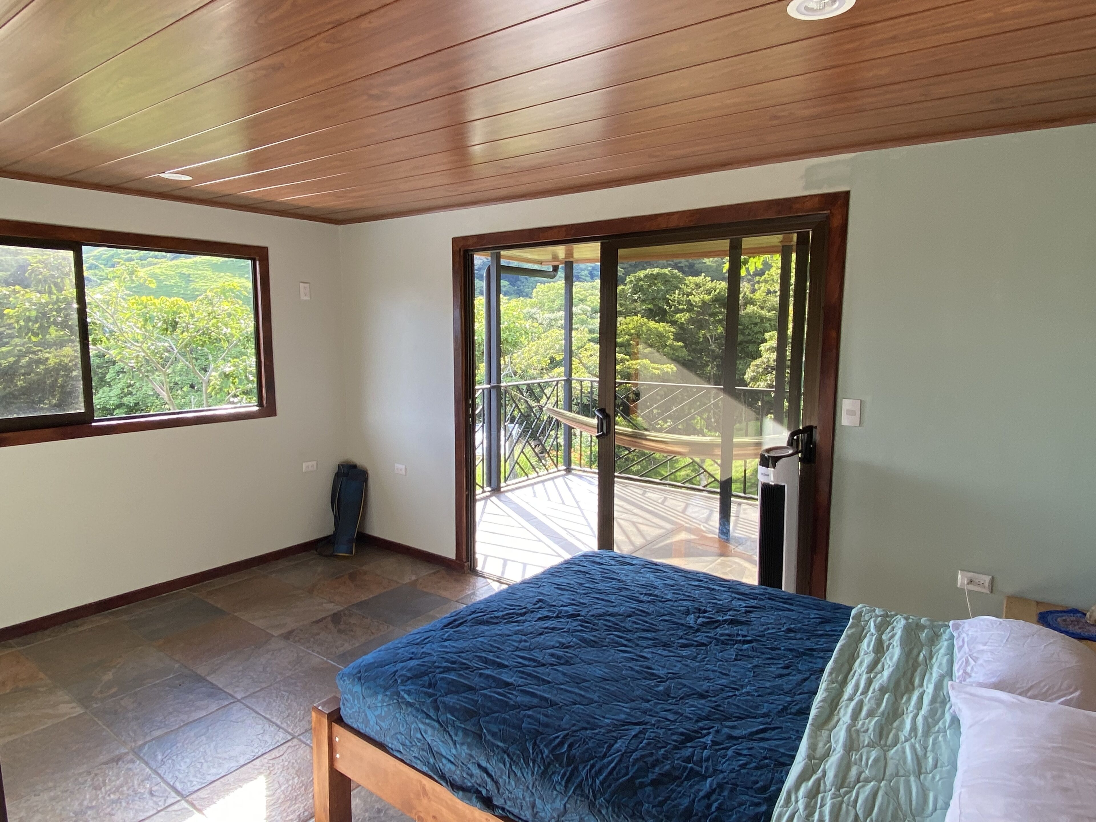 Een slaapkamer met een bed, uitzicht op groen door grote ramen en een balkon met een hekwerk.