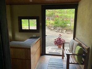 A room with a wooden bench, a small table, a window, and a door leading to an outdoor area with a potted plant and a pile of firewood.