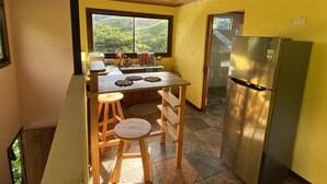 Cocina funcional con encimera de madera, taburetes, refrigerador de acero inoxidable y un amplio ventanal con vistas a la vegetación.