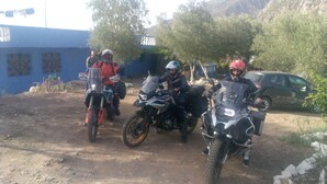 Trois motos sont garées sur un chemin de terre, avec un bâtiment bleu en arrière-plan.