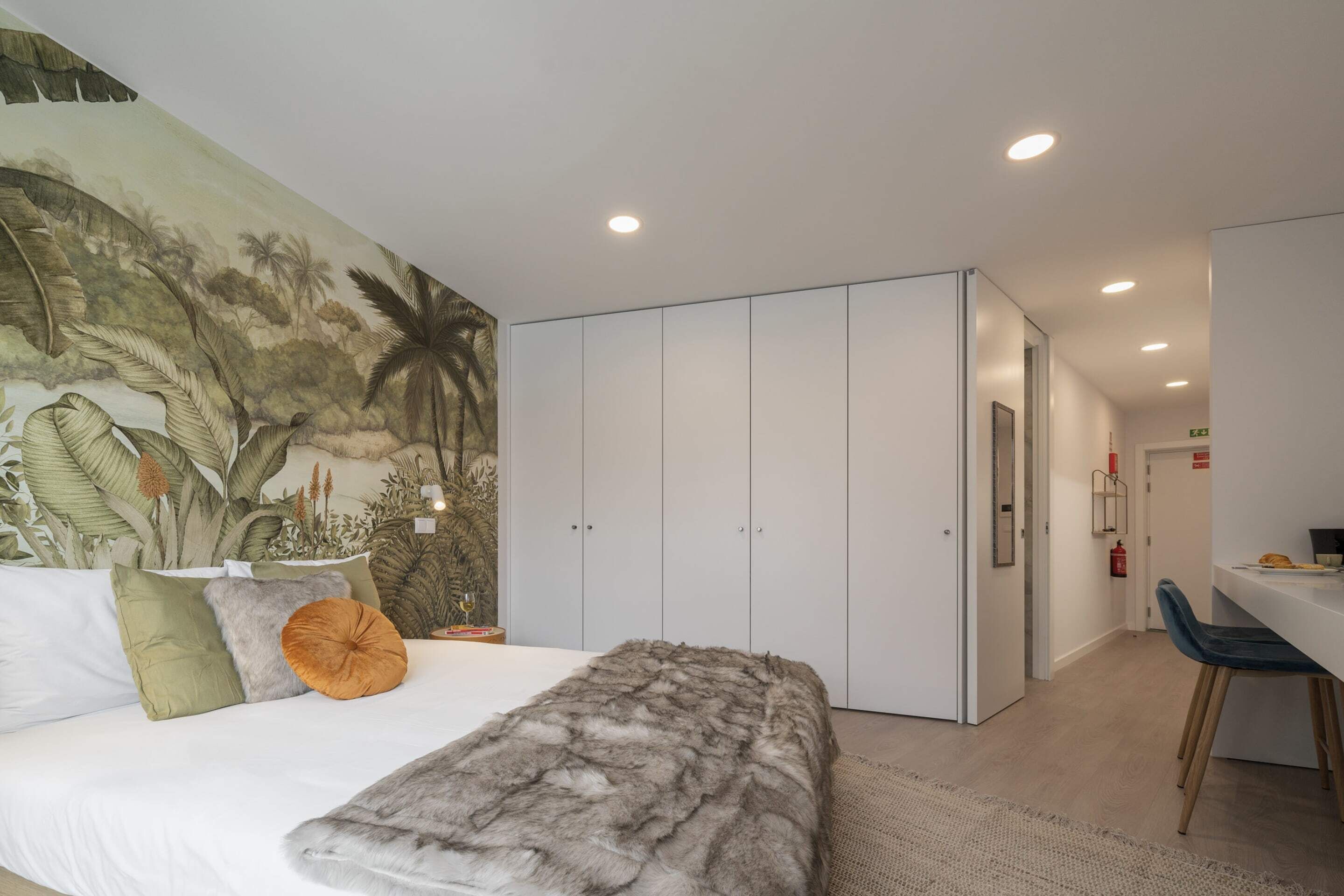 A bedroom with a large bed, a fur throw, a tropical mural, and a white wardrobe.