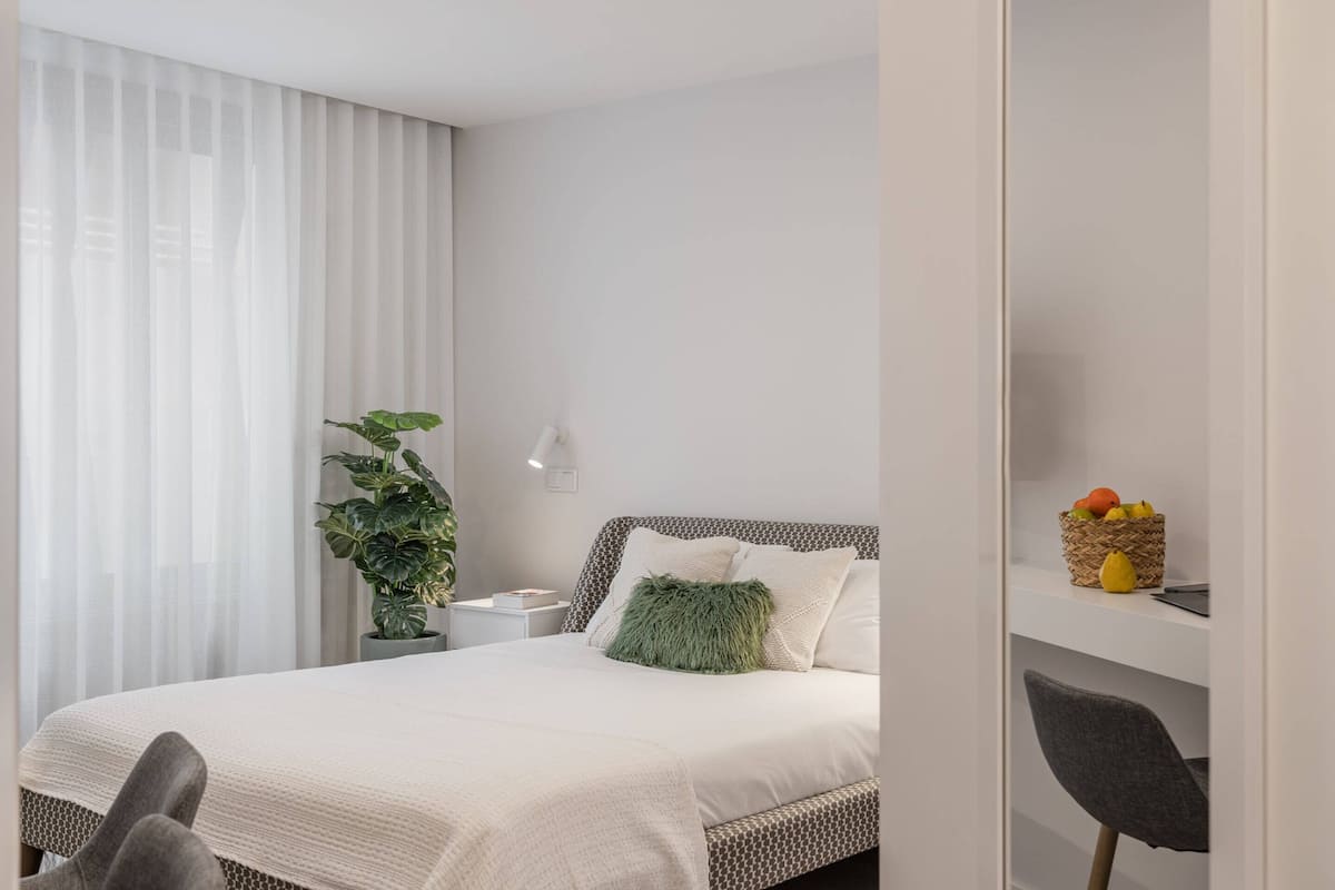 Ein modernes Schlafzimmer mit Bett, Nachttisch, Lampe, Blumentopf, Stuhl und einer Obstschale auf einem Regal.