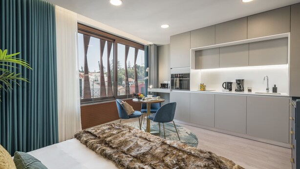 Une chambre moderne avec une grande fenêtre, un plaid douillet en fausse fourrure et un petit coin repas avec des chaises bleues.