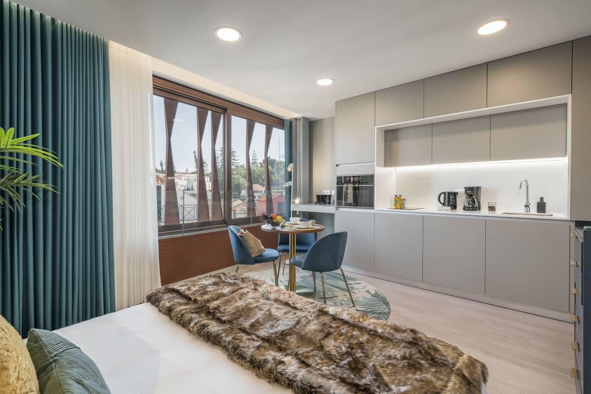 Ein modernes Schlafzimmer mit großem Fenster, einer kuscheligen Felldecke und einer kleinen Essecke mit blauen Stühlen.