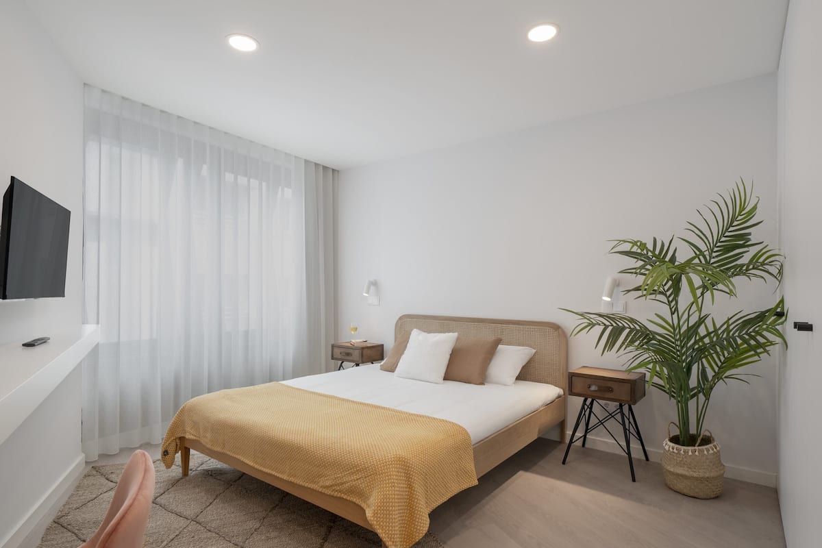 Ein modernes Schlafzimmer mit einem großen Bett, einem an der Wand montierten Fernseher, einem Nachttisch mit Lampe und einem Topf mit Pflanze.