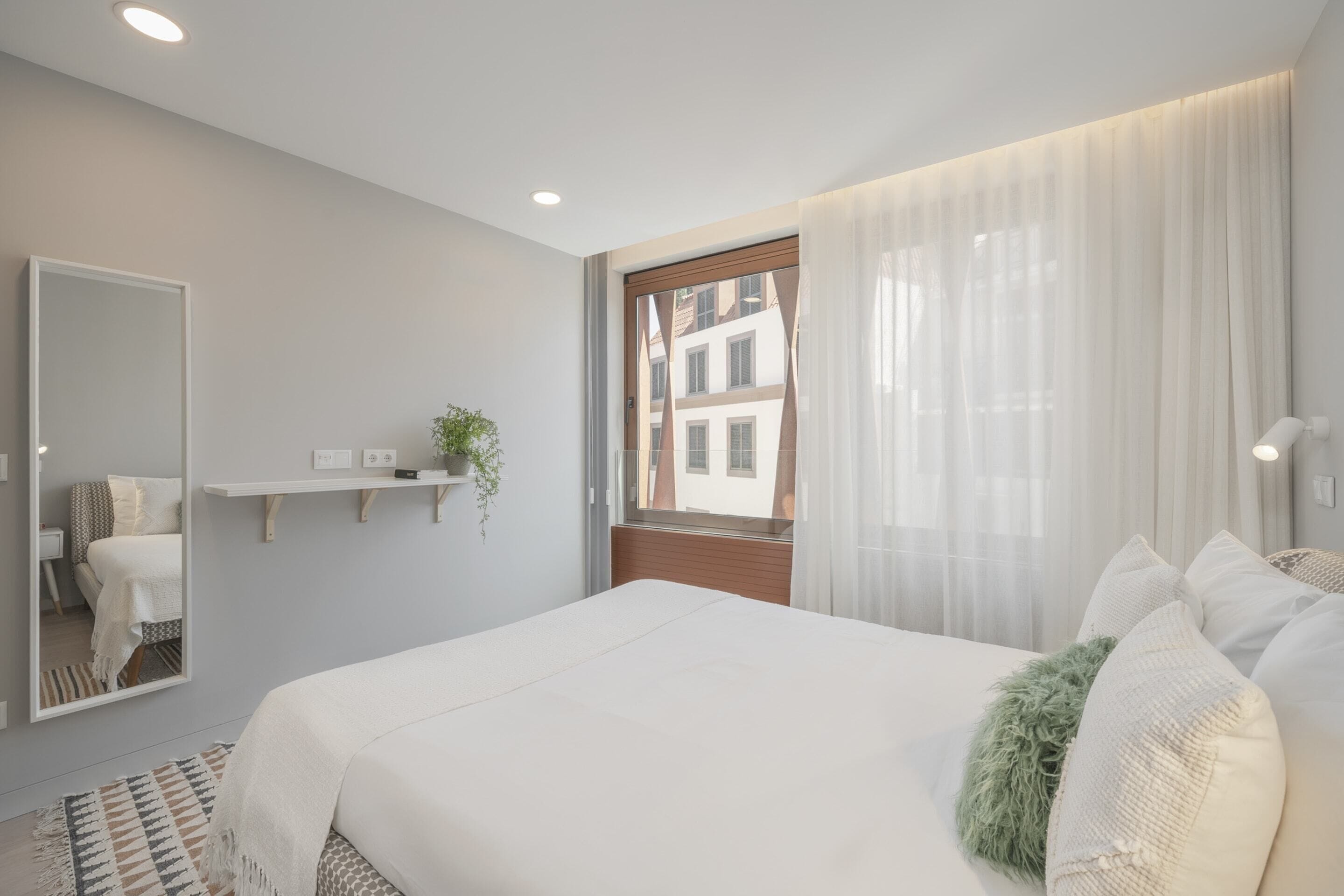 A modern bedroom with a large bed, a mirror, a window with sheer curtains, and a view of buildings outside.
