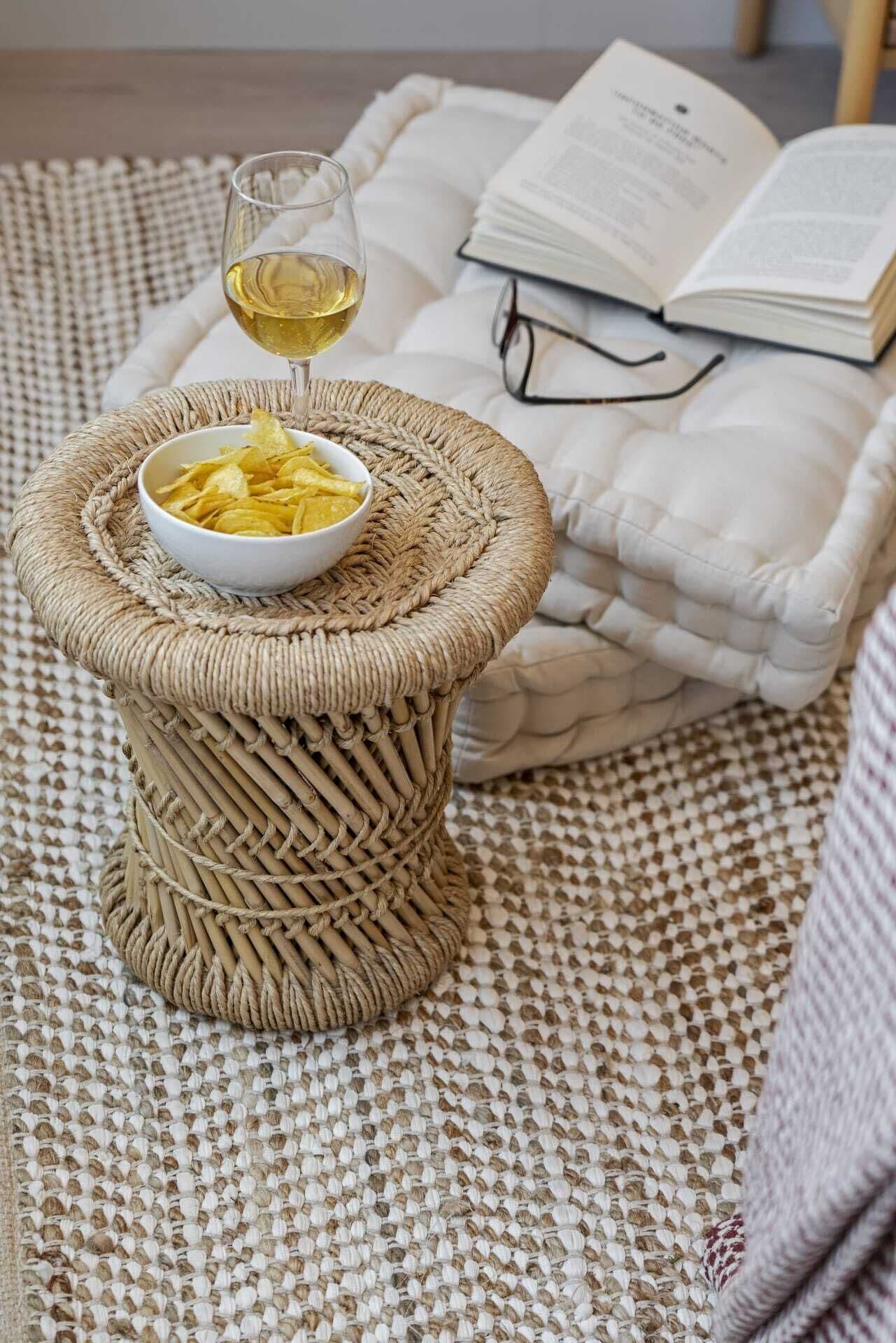 Un tabouret en osier, sur lequel reposent un bol de chips, un verre de vin et un tapis en osier. À côté, sur un lit, on aperçoit un livre et une paire de lunettes.