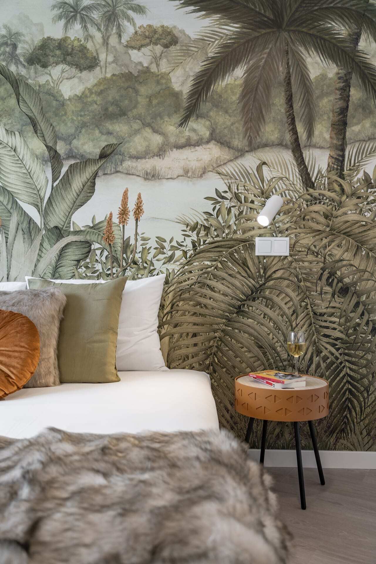 A bedroom with a mural of tropical foliage, a white sofa, and a round wooden side table with a glass of wine.