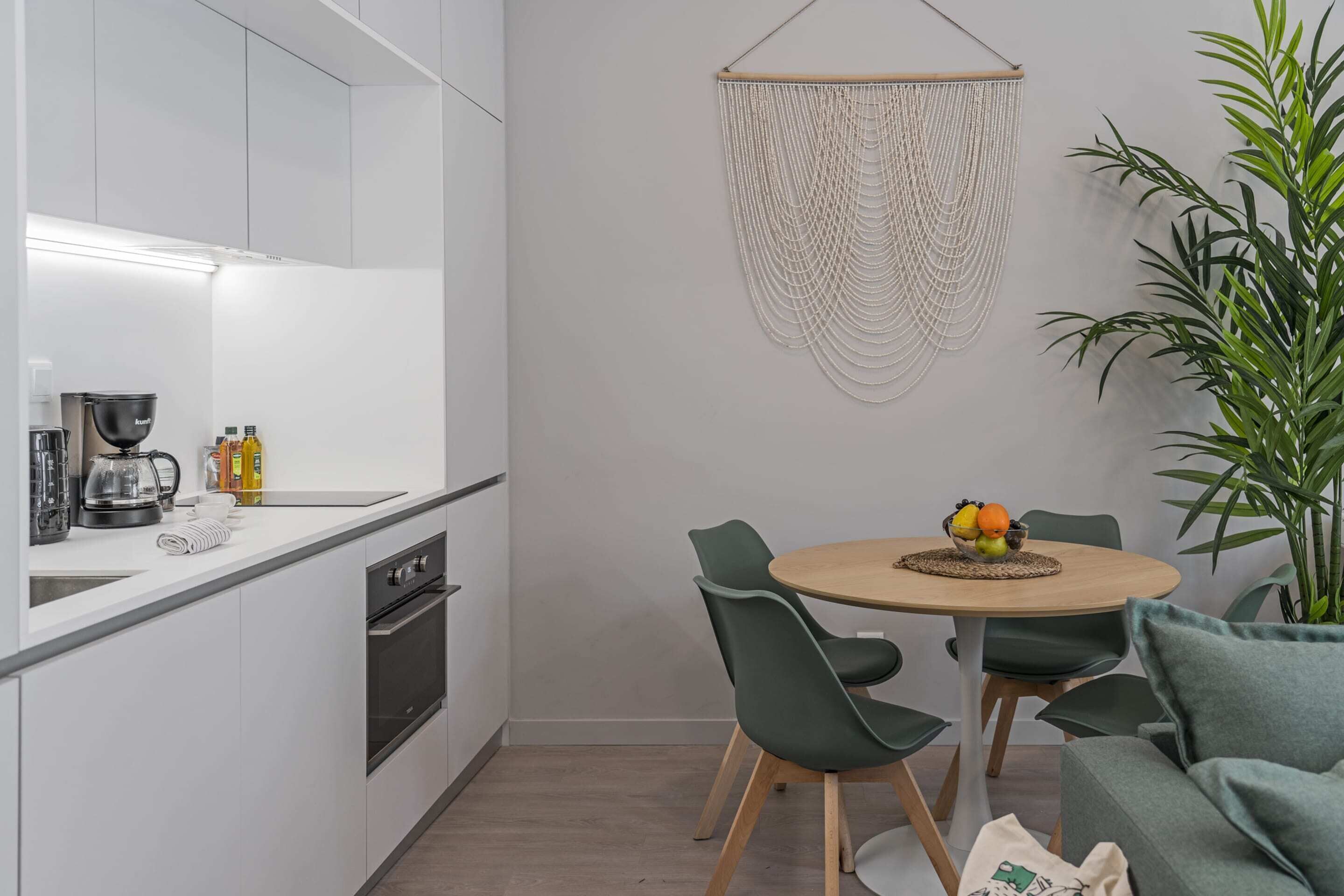 A modern kitchen with white cabinets, a round wooden table with green chairs, and a macrame wall hanging.