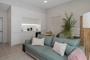 A modern living room with a green sofa, a small round table, and a kitchen area with white cabinets and a bamboo screen.