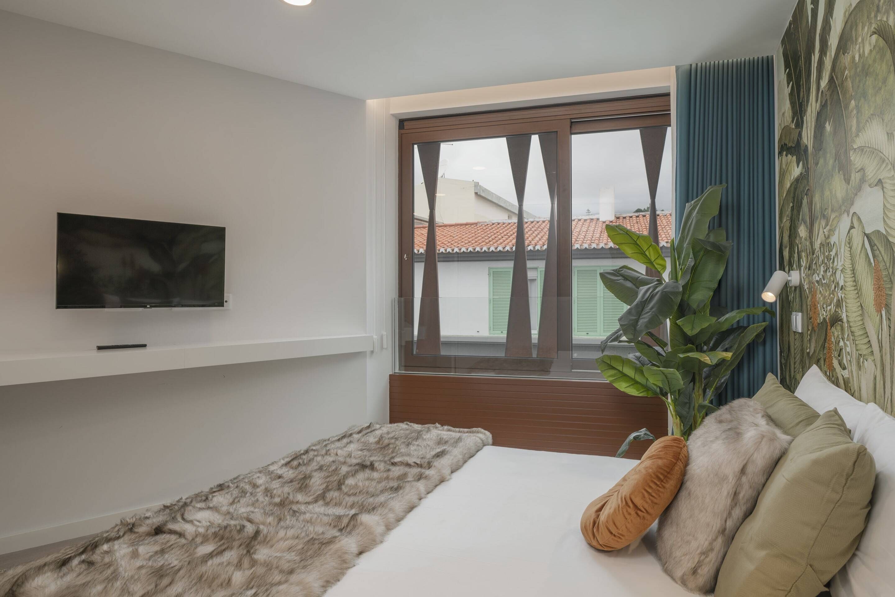 Ein modernes Schlafzimmer mit großem Fenster, einem an der Wand montierten Flachbildfernseher, einem Bett mit flauschiger Decke und einem Blumentopf.
