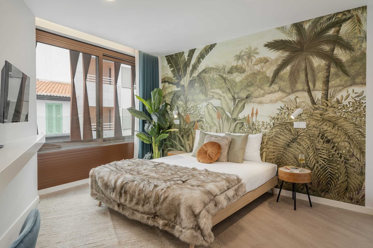 Ein Schlafzimmer mit einem großen Bett, einer Wandmalerei, einem Fenster mit Vorhängen und einem kleinen runden Tisch mit einem Getränk.