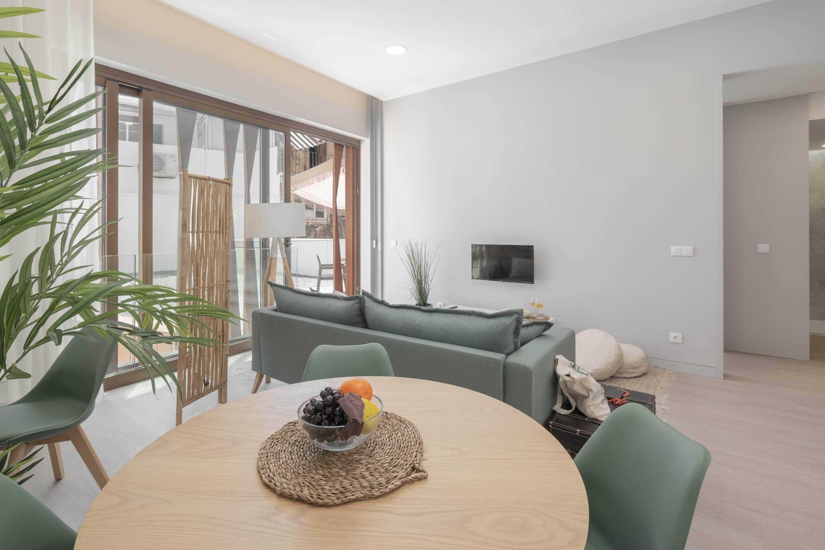 A modern living room with a round wooden table, green chairs, a green sofa, and a TV on the wall.