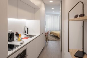 A compact kitchen with a sink, a toaster, and a coffee maker. There is a bed with a yellow bedspread and a chair in the background.