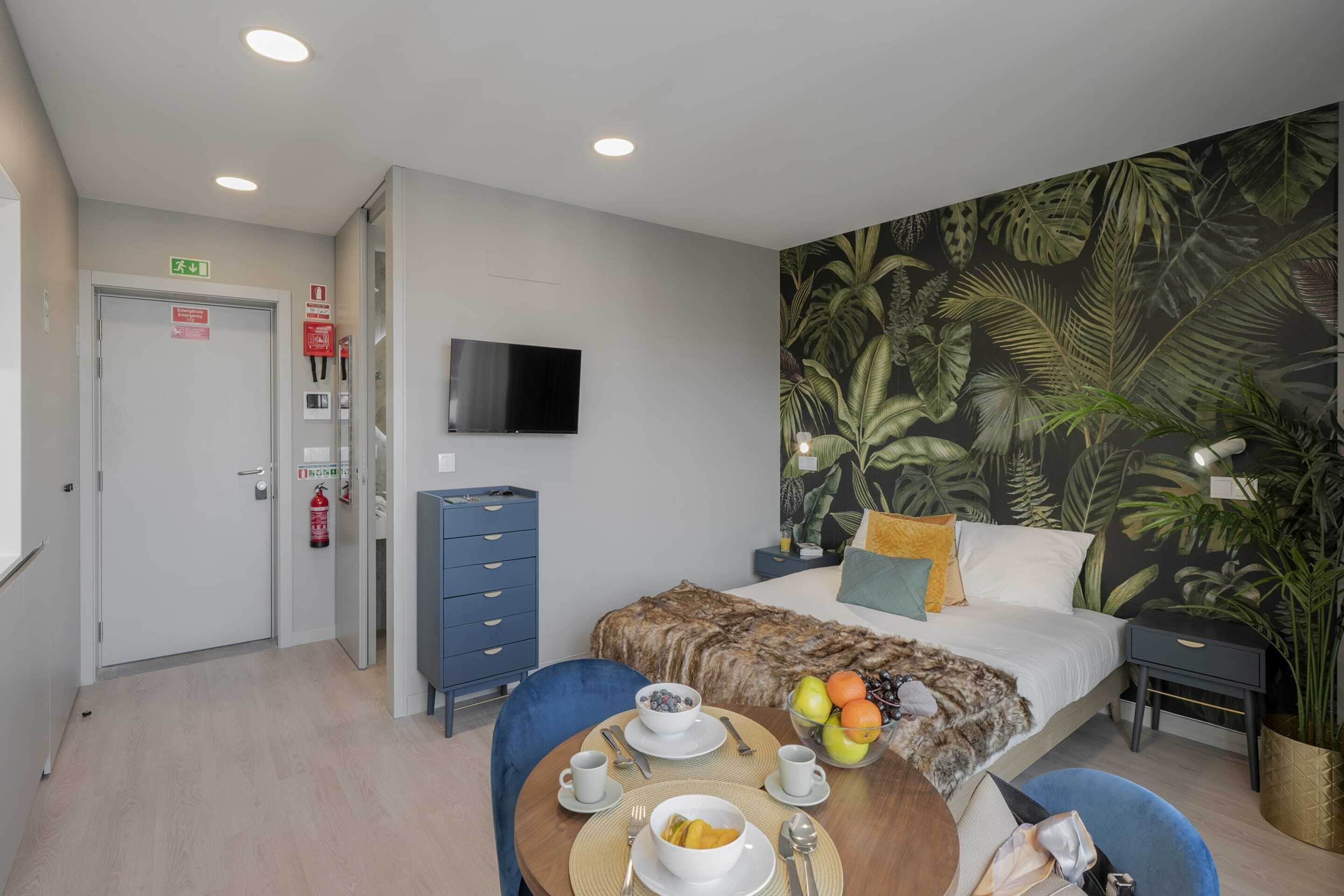 A hotel room with a bed, a TV, a dining area with a table set for breakfast, and a tropical-themed wall mural.