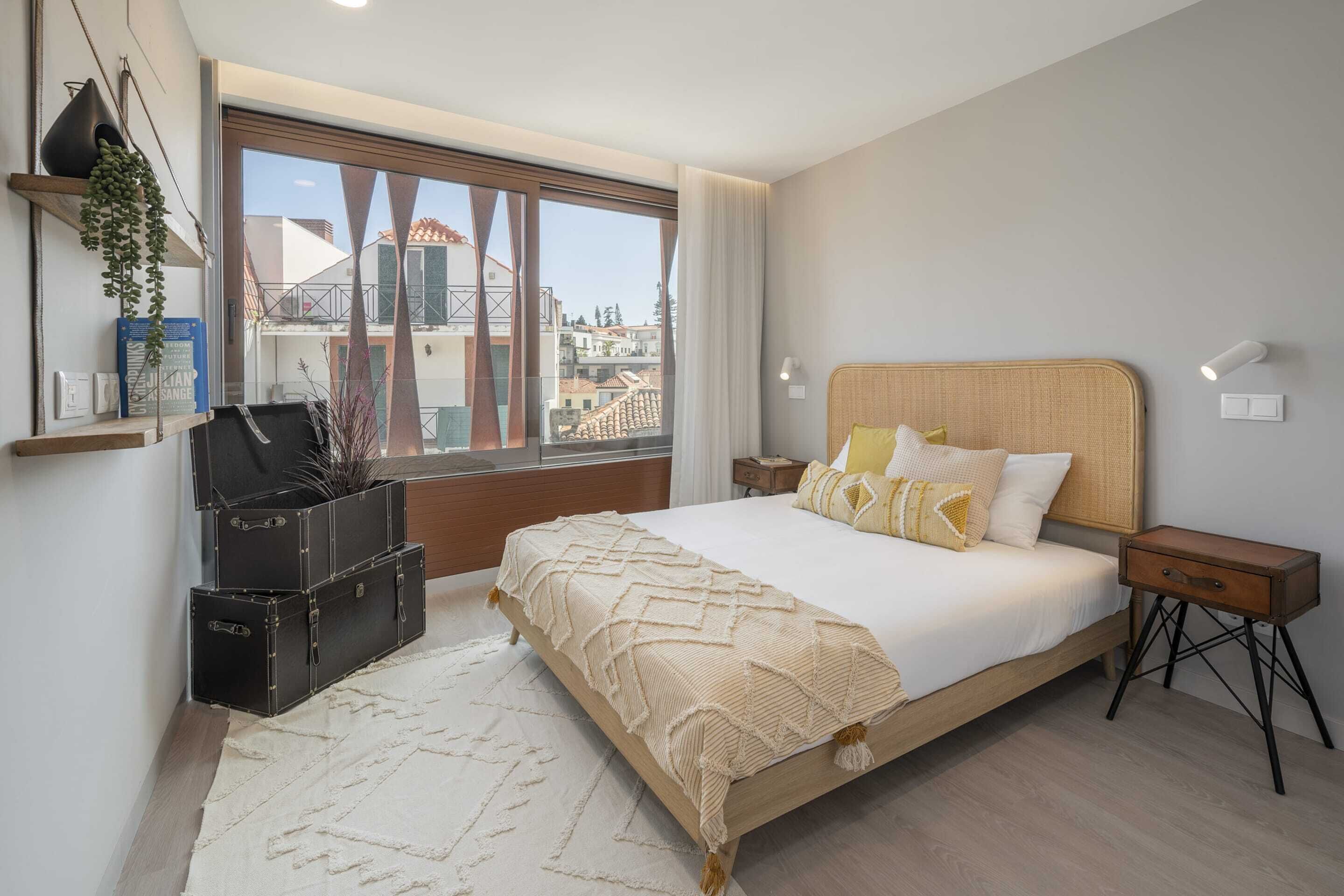 Um quarto com uma cama grande, mesas de cabeceira, uma janela com vista para edifícios e uma planta numa estante.