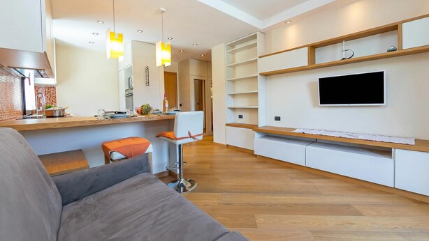 Una sala moderna con un sofá, un comedor con una cocina al fondo y estanterías empotradas con un televisor montado en la pared.