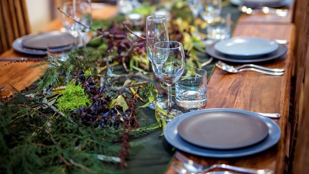 Una larga mesa de madera para comer, dispuesta con platos, vasos y cubiertos, decorada con follaje y elementos decorativos.