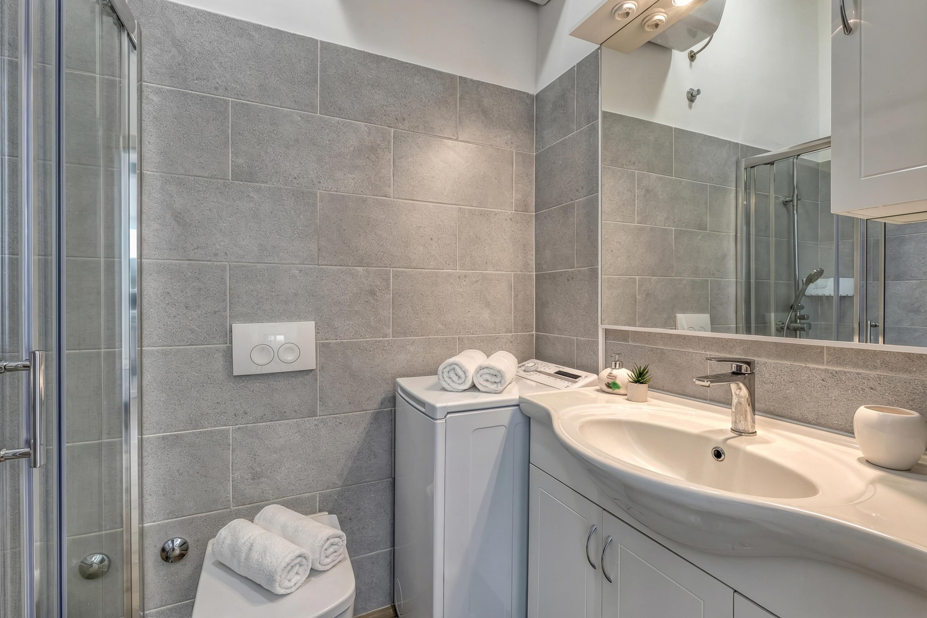 Een moderne badkamer met grijze tegels, een glazen douchecabine, een wit wastafelmeubel en een kast met handdoeken.
