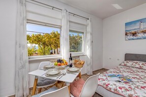 A room with a dining table set for two, a bed with a floral bedspread, and a painting of a lighthouse on the wall. - Pool And Garden Apartment Happy Sound (Rijeka)