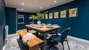 Uma sala de jantar com uma mesa de madeira posta para quatro pessoas, rodeada por cadeiras azuis, um banco e um vaso com flores.
