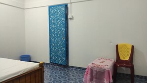 Quarto pequeno e pouco mobiliado, com uma cama, uma porta azul com padrões florais, uma mesa rosa e uma cadeira amarela.
