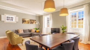 Uma sala de jantar com uma mesa de madeira e seis cadeiras, um sofá, uma televisão e uma janela com cortinas.