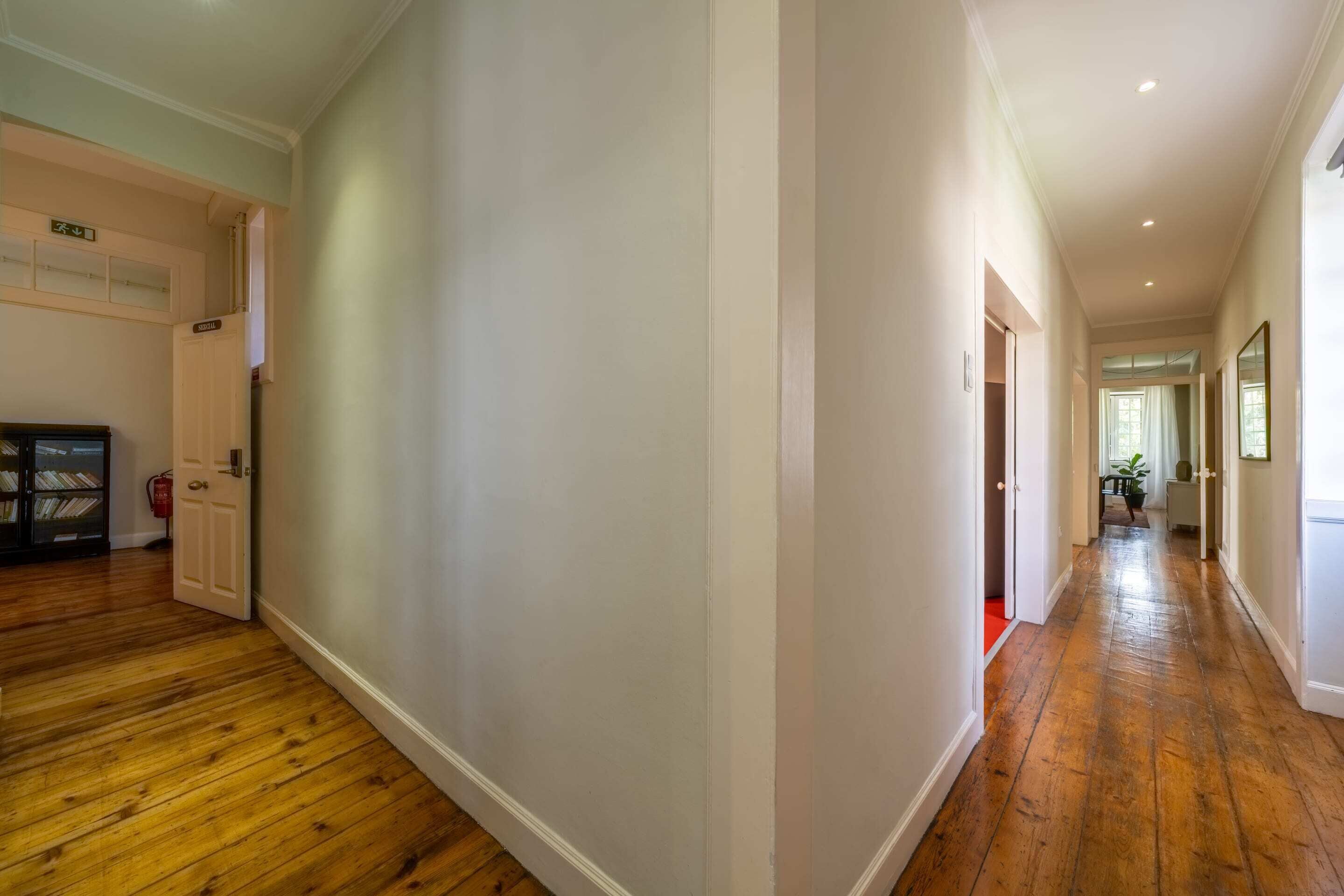 Un couloir doté d’un plancher en bois, de murs blancs et d’une porte donnant sur une pièce où se trouve une moquette rouge.