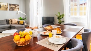 Disposição para o pequeno-almoço com frutas, sumo e pastelaria sobre uma mesa de madeira num quarto claro, com um sofá e televisão ao fundo.