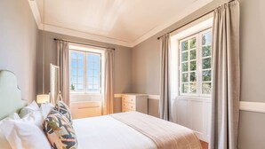 Quarto com cama, mesa de cabeceira, cômoda e duas janelas com cortinas.