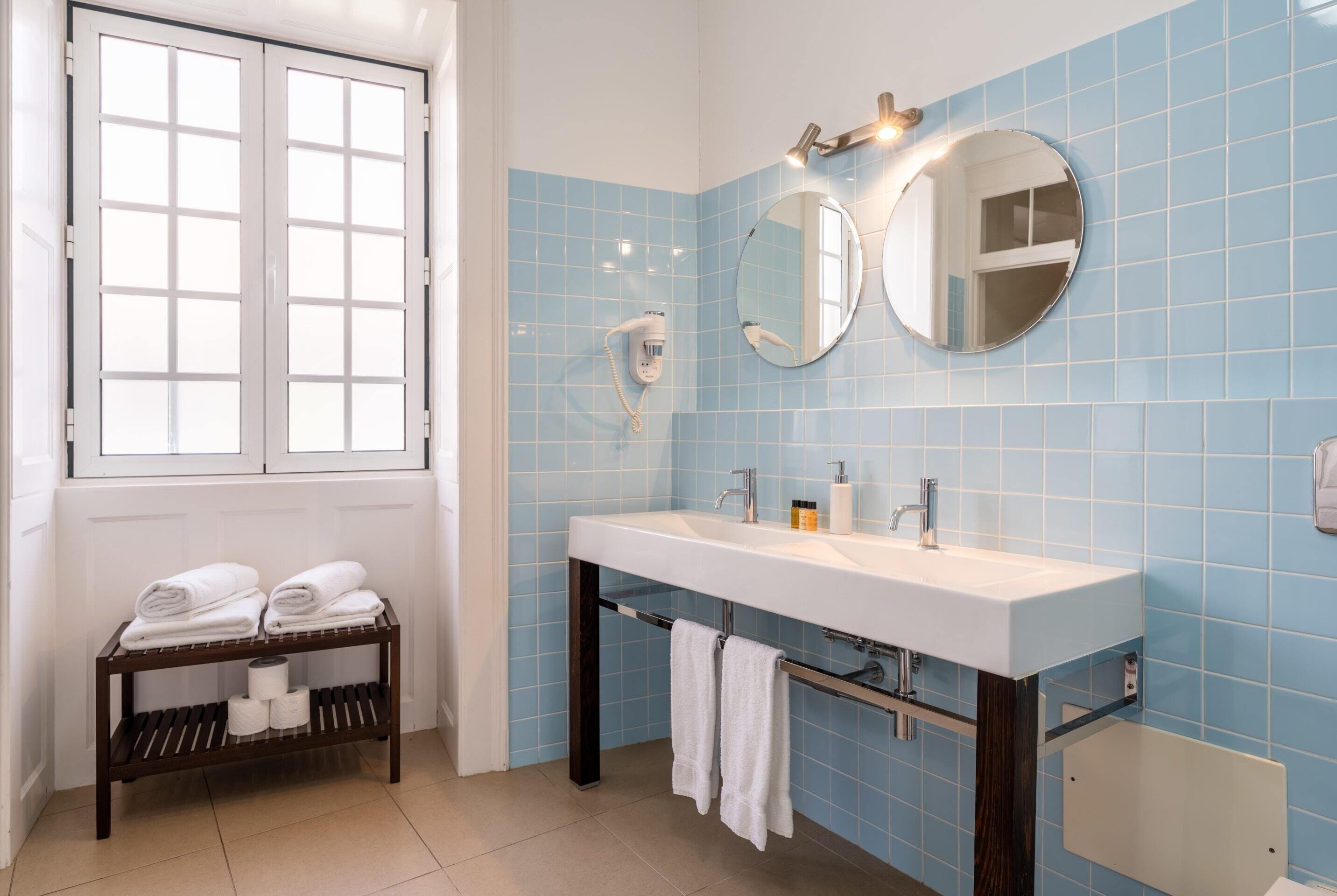 A bathroom with a white sink, two mirrors, a towel rack with towels, a hairdryer, and a window with blinds.
